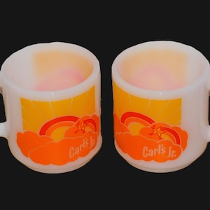 May include: Two white ceramic mugs with a handle. Each mug features a design with a yellow sun, orange clouds, a rainbow, and the text "Carl's Jr." in white. The mugs are set against a black background.