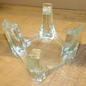May include: A clear glass stand featuring a square base with four thick, upright corner supports. The structure is made of transparent, molded glass with smooth, rounded edges and a hollow center, designed to hold objects securely in place.