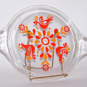May include: A clear glass Pyrex lid featuring a bright orange and yellow floral and bird pattern. The lid has two handles and is presented on a gold-coloured metal stand. The lid is marked with the number "474-012".