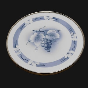 May include: Round white coaster with a blue and silver border. The center features a blue grape cluster and leaves design. The coaster has a silver rim and is set against a black background.