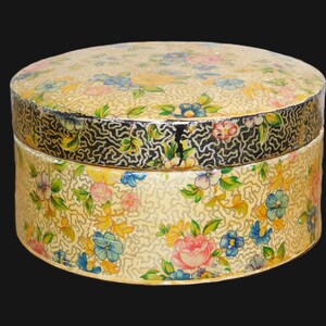 May include: A round, vintage box with a floral pattern. The box is primarily cream-colored with a repeating pattern of pink, blue, and yellow flowers. The lid and base are adorned with a black and silver patterned border.