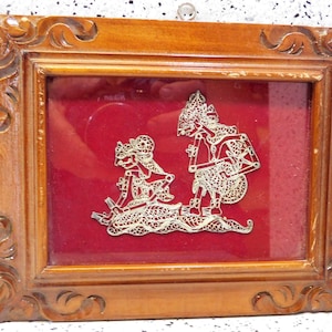 May include: A framed silver cutout of two figures in traditional Indonesian shadow puppet style, set against a red velvet background.