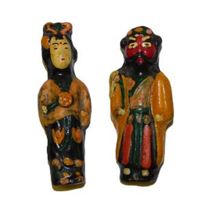 May include: Two hand-painted, vintage Chinese figurines. The figure on the left is a woman with a yellow face, black hair, and an orange and black robe. The figure on the right is a man with a red face, black beard, and an orange and green robe.