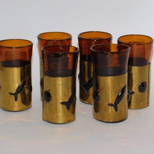 May include: Set of six amber-colored glass tumblers with brass sleeves. The brass sleeves feature black fish-shaped cutouts and small red accents. The glasses have a cylindrical shape and are arranged on a white surface.