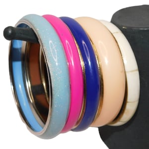 May include: A collection of colorful bangles in various shades of blue, pink, and cream. The bangles are displayed on a black stand, showcasing their glossy and textured surfaces. The set includes a light blue glitter bangle, a pink bangle, and a navy blue bangle.