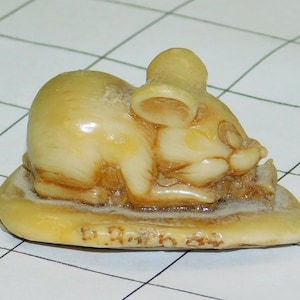 May include: A small, cream-colored figurine of a sleeping animal, possibly a mouse or a rat, resting on a rounded base. The figurine has detailed features and is signed "D. Roth 2004". The base is oval-shaped.