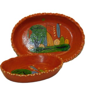 May include: Two terracotta-colored, oval-shaped serving dishes. The larger dish features a hand-painted landscape scene with a green tree, blue and yellow structures, and green grass. The rims have a decorative, textured edge.