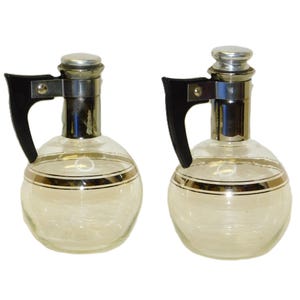 May include: Two clear glass decanters with silver-colored metal accents and black handles. Each decanter has a round body with a gold-colored band around the middle and a silver-colored stopper. The decanters are designed for serving beverages.