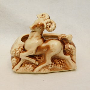 May include: A ceramic planter shaped like a ram with brown and white glaze. The ram is standing on a rocky base with brown and white glaze.