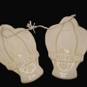 May include: Two off-white ceramic Christmas ornaments. Each ornament features a stylized angel design with the word "NOEL" on a banner. The ornaments are connected by a silver string.