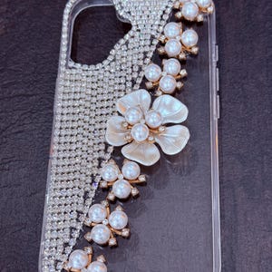 May include: A clear phone case with a sparkly rhinestone design and a pearl and flower embellishment.