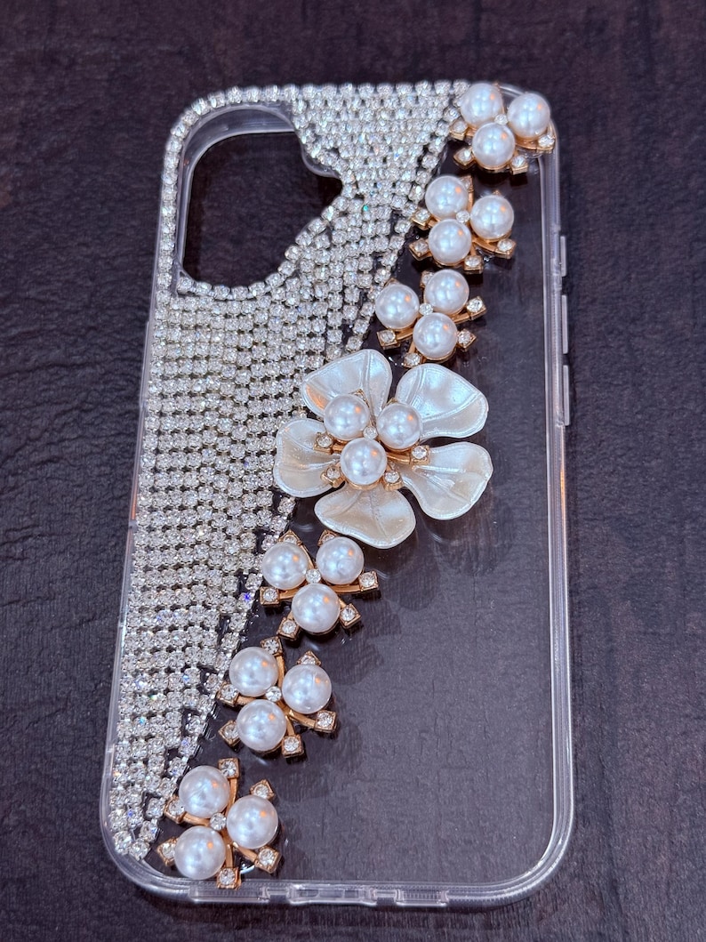May include: A clear phone case with a sparkly rhinestone design and a pearl and flower embellishment.