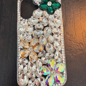 May include: A clear phone case decorated with rhinestones and three flower-shaped embellishments. The flowers are green, gold, and iridescent.