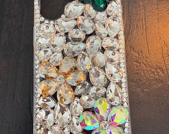 May include: A clear phone case decorated with rhinestones and three flower-shaped embellishments. The flowers are green, gold, and iridescent.