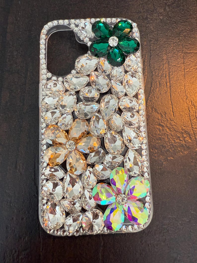 May include: A clear phone case decorated with rhinestones and three flower-shaped embellishments. The flowers are green, gold, and iridescent.