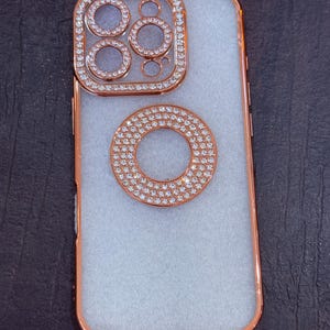 May include: A white phone case with a rose gold trim and a diamond-encrusted ring around the camera lens.