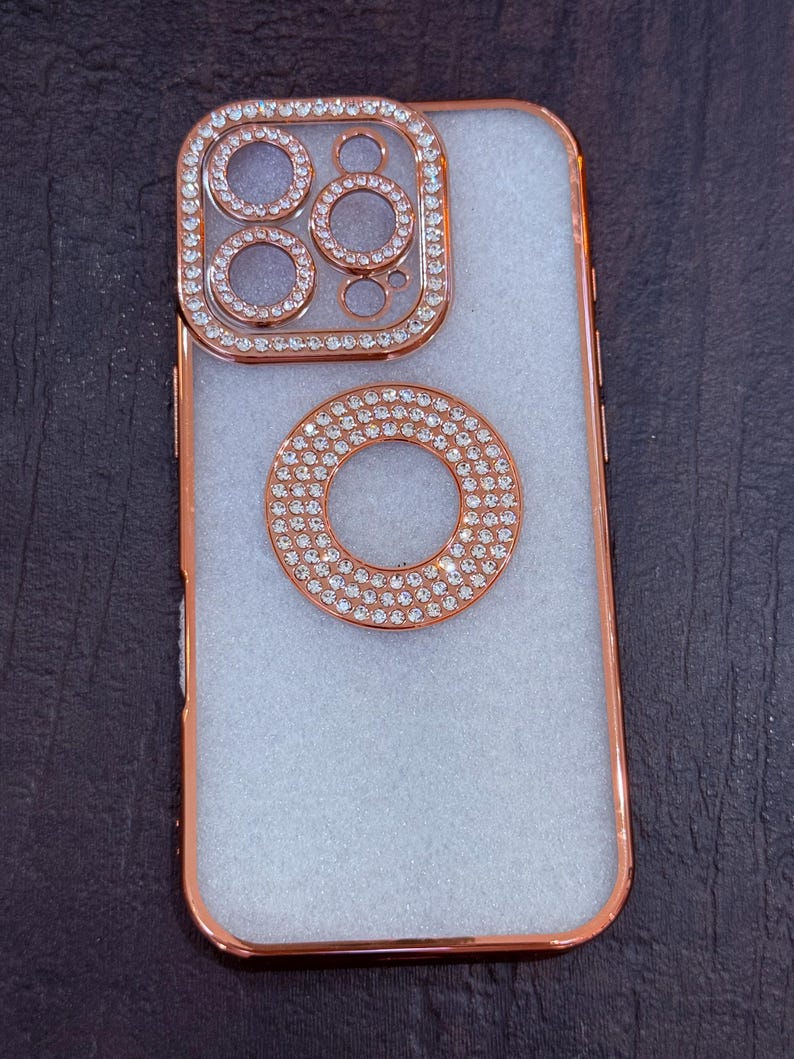 May include: A white phone case with a rose gold trim and a diamond-encrusted ring around the camera lens.
