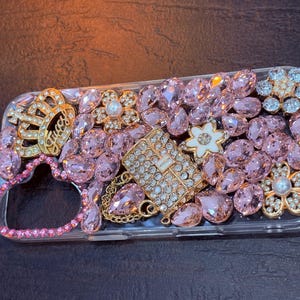 May include: A clear phone case decorated with pink, white, and gold rhinestones. The case features a crown, a flower, and a small purse design.