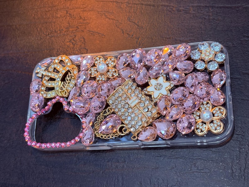 May include: A clear phone case decorated with pink, white, and gold rhinestones. The case features a crown, a flower, and a small purse design.