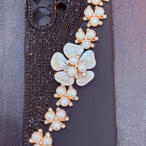 May include: Black phone case with a pearl and rhinestone design. The design features a large white flower with a pearl centre and a row of pearl and rhinestone bows.