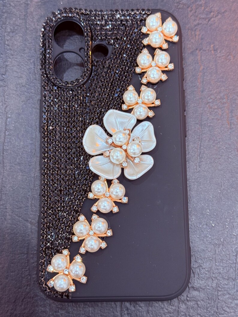 May include: Black phone case with a pearl and rhinestone design. The design features a large white flower with a pearl centre and a row of pearl and rhinestone bows.