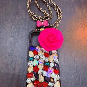 May include: A black phone case with a gold chain, a pink bow, a pink flower, and a red lip design. The case is covered in rhinestones of various colours.