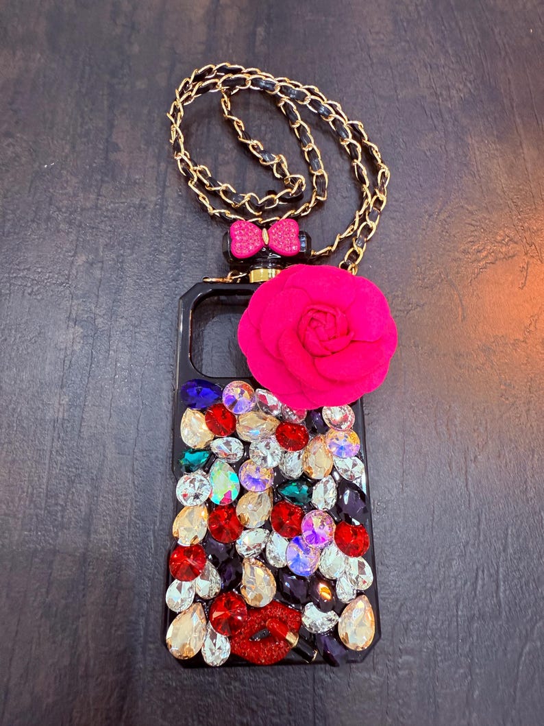 May include: A black phone case with a gold chain, a pink bow, a pink flower, and a red lip design. The case is covered in rhinestones of various colours.