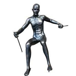 May include: A silver-colored metal sculpture of a person in a seated position, holding two sticks in their hands. The sculpture is posed as if the person is ice skating.
