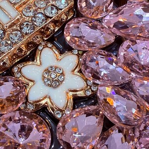 May include: A close-up of a gold-toned flower-shaped brooch with white enamel and clear rhinestones, surrounded by pink and clear glass rhinestones.