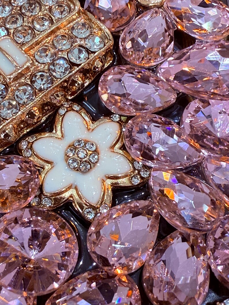 May include: A close-up of a gold-toned flower-shaped brooch with white enamel and clear rhinestones, surrounded by pink and clear glass rhinestones.
