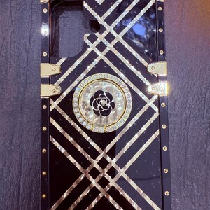 May include: Black phone case with a gold geometric pattern and a rhinestone-encrusted ring holder featuring a black flower.