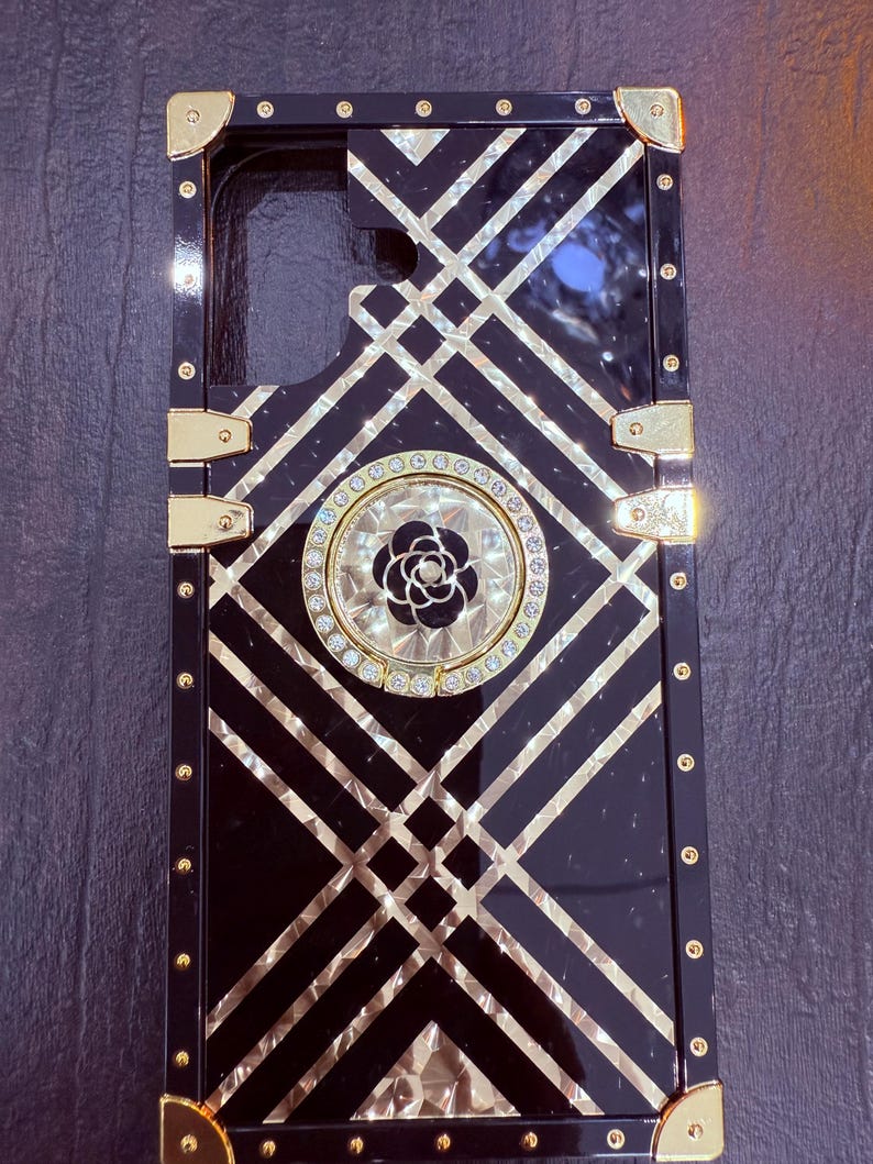 May include: Black phone case with a gold geometric pattern and a rhinestone-encrusted ring holder featuring a black flower.