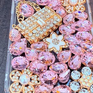 May include: A clear phone case decorated with pink and white rhinestones, a gold chain, and a gold purse charm. The case is covered in a variety of shapes and sizes of rhinestones.