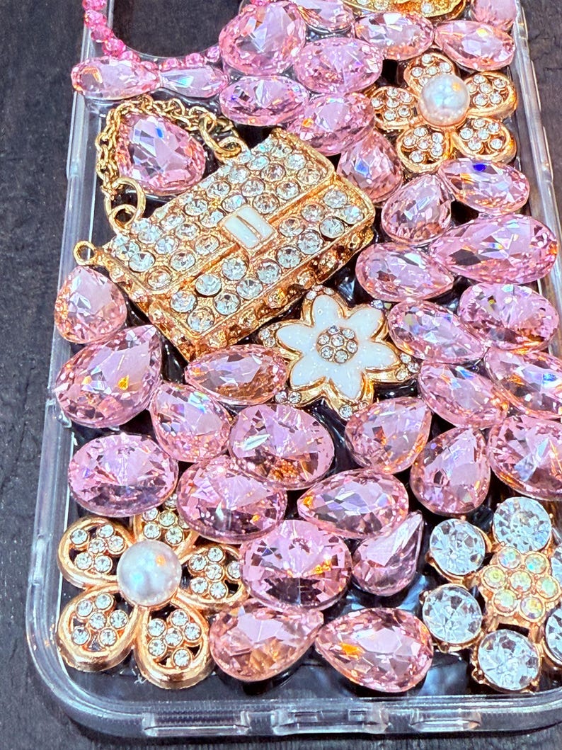 May include: A clear phone case decorated with pink and white rhinestones, a gold chain, and a gold purse charm. The case is covered in a variety of shapes and sizes of rhinestones.