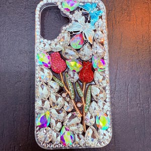 May include: A clear phone case with a floral design. The case is covered in rhinestones and features two red flowers, a butterfly, and green leaves.