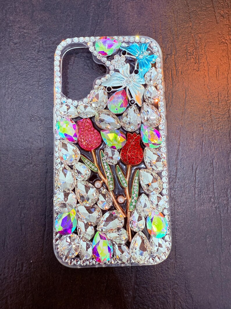 May include: A clear phone case with a floral design. The case is covered in rhinestones and features two red flowers, a butterfly, and green leaves.