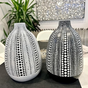 May include: Two decorative vases with a black and white polka dot pattern. One vase is white with black dots, and the other is black with white dots. The vases have a rounded shape and a narrow neck. They are placed on a black surface.