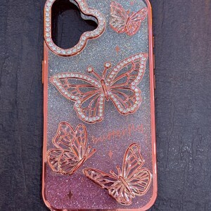 May include: A rose gold phone case with a glittery pink and silver gradient background. The case features four butterfly designs with rhinestones. The text "butter fly" is printed on the case.