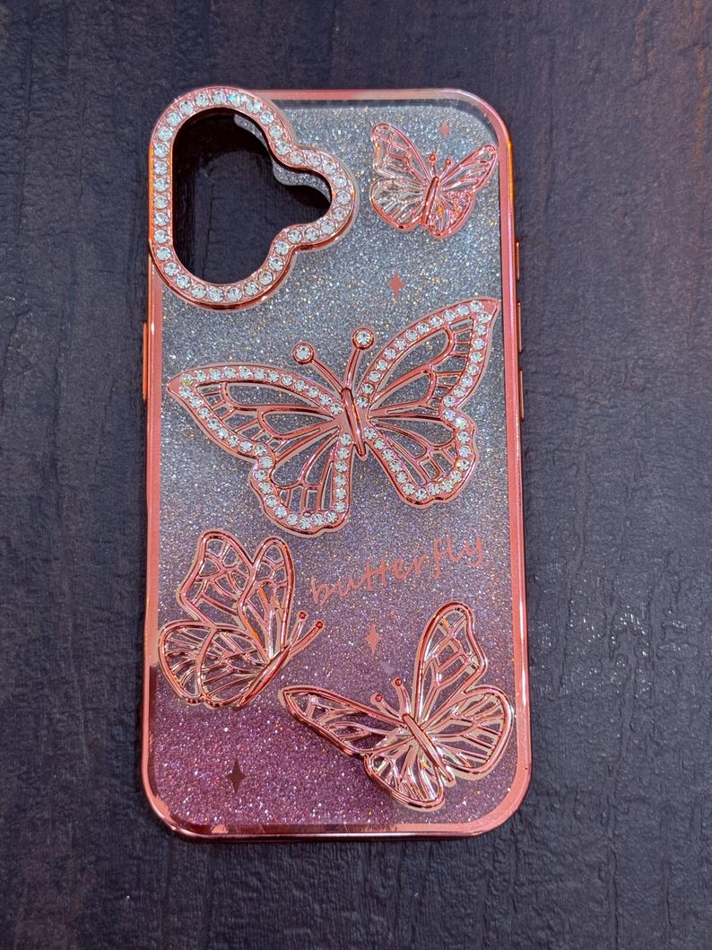 May include: A rose gold phone case with a glittery pink and silver gradient background. The case features four butterfly designs with rhinestones. The text "butter fly" is printed on the case.