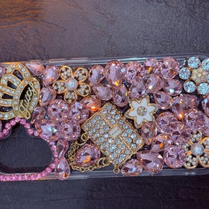 May include: A clear phone case decorated with pink, gold, and white rhinestones. The case features a crown, a heart, and a small purse design.