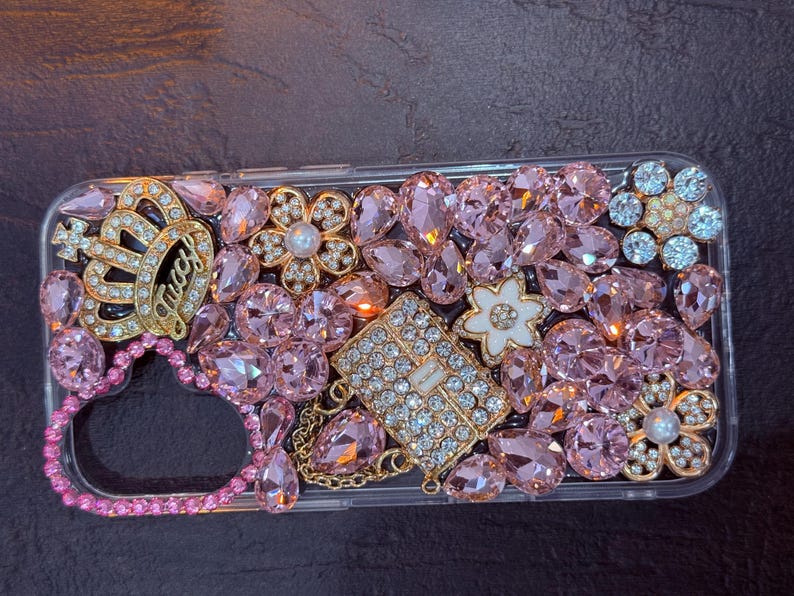 May include: A clear phone case decorated with pink, gold, and white rhinestones. The case features a crown, a heart, and a small purse design.
