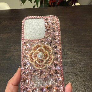May include: A clear phone case with a pink and gold flower design. The case is covered in rhinestones and has a pearl in the centre of the flower.