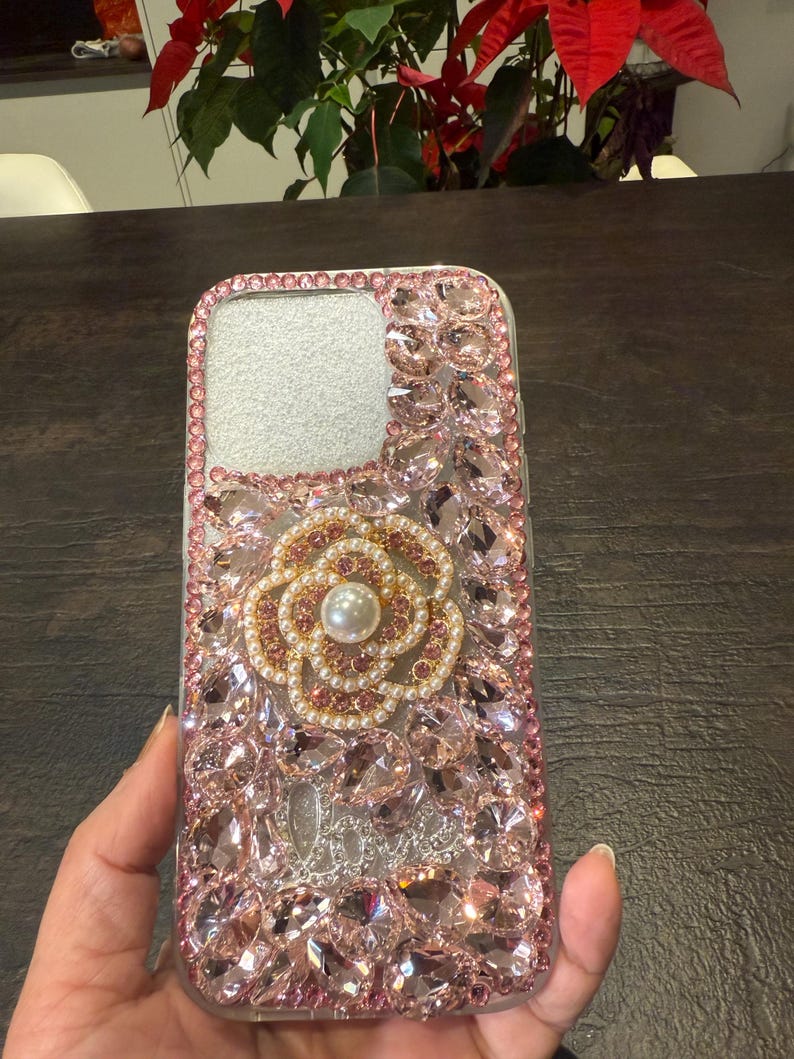 May include: A clear phone case with a pink and gold flower design. The case is covered in rhinestones and has a pearl in the centre of the flower.