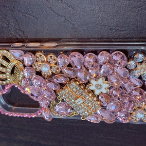 May include: A clear phone case decorated with pink and gold rhinestones, including a crown, a flower, and a small purse.