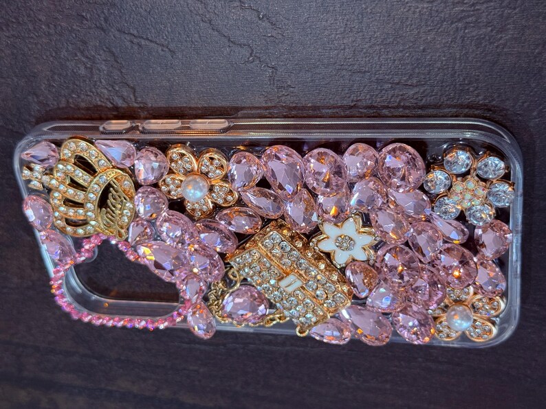 May include: A clear phone case decorated with pink and gold rhinestones, including a crown, a flower, and a small purse.