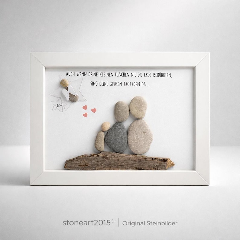 May include: A framed artwork featuring a family crafted from smooth stones, resting on a piece of driftwood. The artwork includes a small angel figure and text in German. The frame is white.