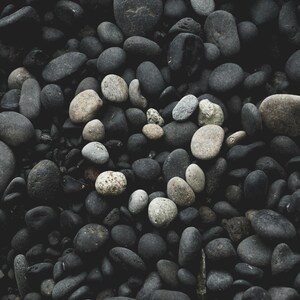 May include: A collection of smooth, dark gray and black river rocks. A heart shape is formed in the center with lighter gray and tan stones. The rocks vary in size and shape, creating a natural, textured pattern.