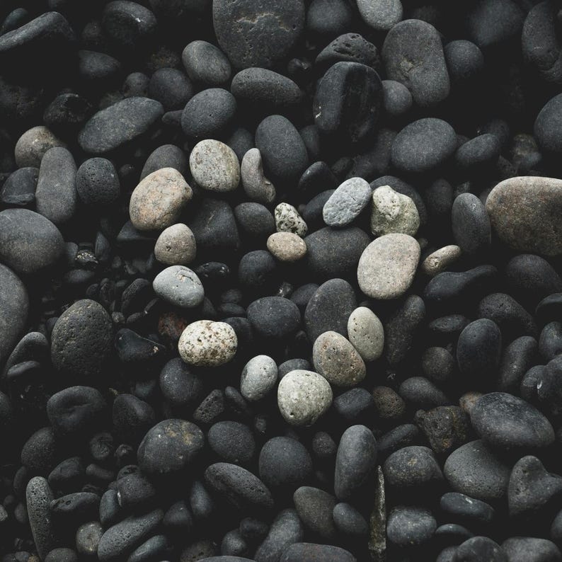 May include: A collection of smooth, dark gray and black river rocks. A heart shape is formed in the center with lighter gray and tan stones. The rocks vary in size and shape, creating a natural, textured pattern.