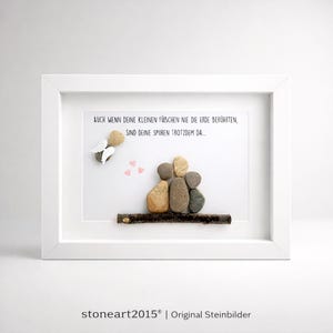 May include: A framed artwork featuring a family of four depicted with smooth stones on a wooden branch. Above, an angel made of stones and three pink hearts are displayed. The text reads, "AUCH WENN DEINE KLEINEN F&Uuml;BCHEN NIE DIE ERDE BER&Uuml;HRTEN, SIND DEINE SPUREN TROTZDEM DA..."