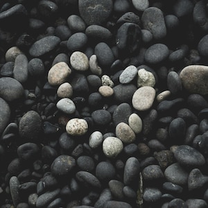 May include: A collection of smooth, rounded river stones in various shades of charcoal, slate gray, and black. A selection of lighter, off-white and beige pebbles are arranged in the center to form a distinct heart shape against the dark background.